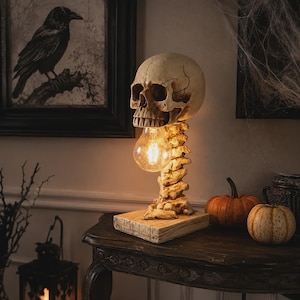 May include: A table lamp featuring a human skull resting atop a spinal column base, which is mounted on a rectangular wooden block. The lamp includes a single glowing light bulb positioned within the open jaw of the skull, casting warm light.