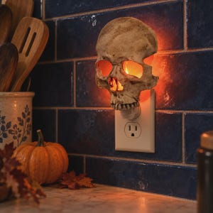 May include: A beige, textured skull-shaped night light plugged into a wall outlet. The skull features hollow eye sockets and a nasal cavity that emit a warm, orange glow from an internal light source, casting light onto the surrounding wall.