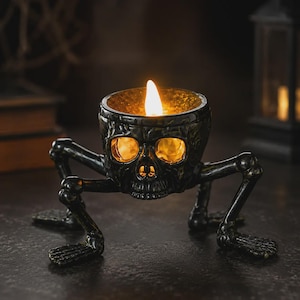 May include: A black ceramic candle holder shaped like a human skull with four skeletal legs extending outward. The top of the skull features a circular opening containing a glowing, warm-toned flame that illuminates the eye sockets and nasal cavity.
