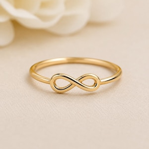 May include: A polished gold ring featuring a central infinity symbol design. The band is thin and circular, connecting to the horizontal figure-eight shape at the front. The metal has a smooth, reflective surface with a warm yellow gold tone.