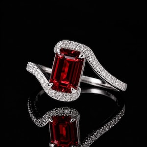 May include: A silver-toned ring featuring a large, rectangular, emerald-cut deep red gemstone set in the center. The band and the setting surrounding the central stone are encrusted with small, clear, sparkling white accent stones.