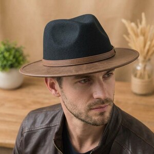 May include: A dark navy blue fedora hat featuring a wide, stiff brim and a smooth, light brown faux leather band encircling the crown. The hat has a structured, pinched top design and a clean, minimalist aesthetic with a flat, wide brim.