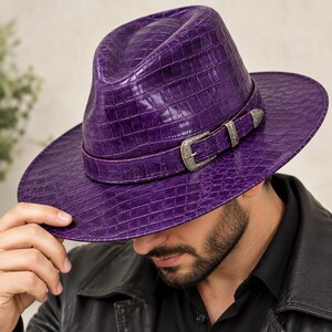 Könnte beinhalten: Ein leuchtend violetter Fedora-Hut mit einer strukturierten Krokodilleder-Optik. Der Hut ist mit einem passenden violetten Band und einer dekorativen, silberfarbenen Metallschnalle mit filigranen Gravuren an der Seite versehen.