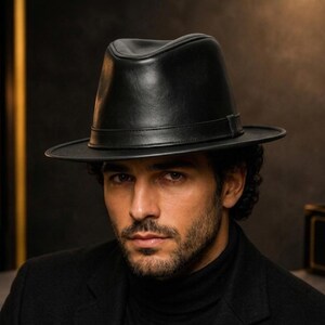 Peut inclure: Un chapeau fedora noir avec une finition lisse et mate et une calotte structurée. Le chapeau présente une bande noire assortie entourant la base de la calotte et un bord plat de largeur moyenne qui s'étend horizontalement sur tout le pourtour.