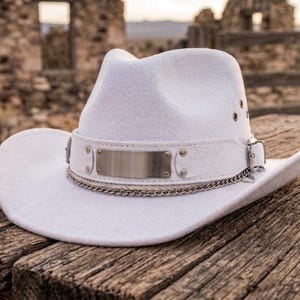 May include: A white cowboy hat featuring a wide brim and a pinched crown. The hat is adorned with a white leather band, a rectangular silver-toned metal plate, silver rivets, a metallic chain detail, and a small silver buckle on the side.