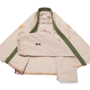 May include: A beige martial arts gi featuring an olive green collar and trim. The interior lining displays a patterned design. Black Japanese characters are embroidered on the front panel. The garment includes long sleeves and reinforced stitching.