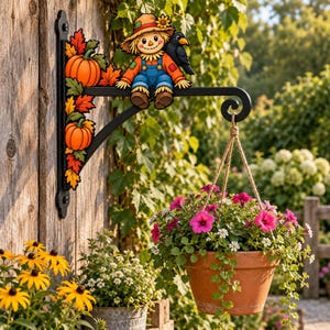 May include: A black metal wall-mounted plant hanger featuring a decorative scarecrow, a black crow, and orange pumpkins with autumn leaves. A terracotta pot containing pink flowers and green foliage hangs from the hook by a jute rope.