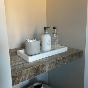 May include: A white rectangular tray holding a grey cylindrical container filled with white cotton swabs, an opaque white pump bottle labelled Molton Brown, and a clear pump bottle labelled Molton Brown, all arranged on a rustic wooden shelf.