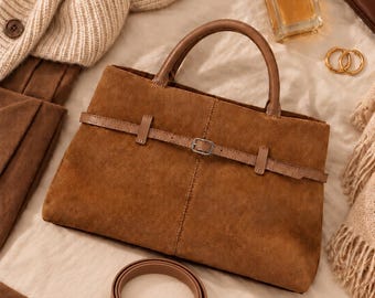 Puede incluir: Un bolso de mano de ante marrón con una correa horizontal con hebilla de metal plateado en la parte delantera, dos asas superiores y una correa de hombro a juego extraíble con clips de metal plateado, de diseño estructurado.
