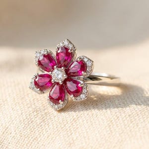 Könnte beinhalten: Ein silberfarbener Ring mit einem blumenförmigen Mittelstück. Die Blume besteht aus sechs tiefroten, birnenförmigen Edelsteinen als Blütenblätter, einem runden weißen Edelstein in der Mitte und kleinen weißen Akzentsteinen am Rand.