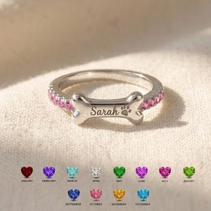 May include: A silver-toned ring featuring a central bone-shaped plaque engraved with the name Sarah and a small paw print. The band is accented with pink gemstones along the sides. Below the ring, twelve colored heart icons represent birth months.