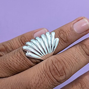 May include: A silver-toned ring featuring a fan-shaped design that resembles a seashell. The surface of the ring is smooth and reflective, with distinct vertical ridges radiating from the base to the curved, scalloped top edge of the piece.