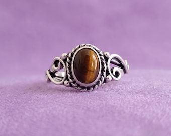 May include: A silver ring featuring an oval-shaped tiger's eye gemstone set in a twisted rope-style bezel. The band is decorated with intricate silver scrollwork and small spherical accents on both sides of the central stone setting.