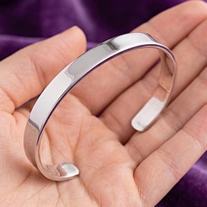May include: A polished silver-tone cuff bracelet with a smooth, reflective surface and a wide, open-ended band design. The metal has a high-shine finish that catches the light, creating bright highlights along the curved edges of the bracelet.