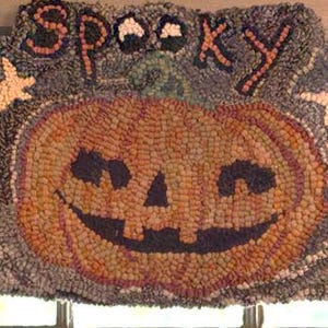 Op de afbeelding: Een Halloween-tapijt met een lachende pompoen, twee zwarte katten en het woord "Spooky". Het tapijt heeft een grijze achtergrond met bruine, oranje en zwarte details. Twee witte sterren flankeren de pompoen.