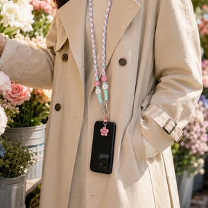 May include: A phone lanyard featuring a braided strap with a repeating pattern of small pink and white flowers, connected to a black smartphone case by a silver metal clasp and a small pink flower charm attached to the bottom of the case.