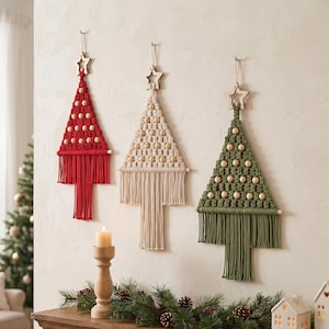 May include: Three macrame wall hangings shaped like Christmas trees in red, cream, and olive green. Each tree features wooden beads, a wooden star topper, and a fringed base. The hangings are suspended by string against a neutral wall.
