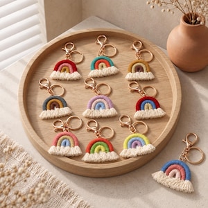 Puede incluir: Una colección de diez llaveros de arcoíris de macramé con varias combinaciones de colores de cordón de algodón, cada uno unido a un cierre de mosquetón y una anilla de metal dorado. Los arcoíris tienen una base con flecos sobre una bandeja de madera.