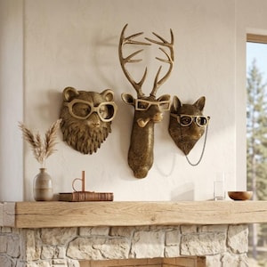 Può includere: Tre sculture da parete a forma di testa di animale con finitura color bronzo, raffiguranti un orso, un cervo con corna e una volpe. Ogni animale indossa occhiali con montatura dorata, il cervo ha una pipa in bocca e la volpe una catenella.