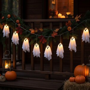 May include: A string of LED lights featuring white fabric ghost covers with black eyes and mouths, draped across a wooden railing decorated with evergreen branches, autumn leaves, and small pumpkins, flanked by two metal lanterns with lit candles.