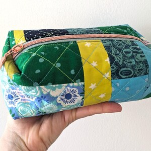 Op de afbeelding: Een rechthoekig, doorgestikt etui met ritssluiting en een patchworkontwerp van diverse stoffen in groen-, geel- en blauwtinten. De patronen op de stof bevatten bloemmotieven, kleine sterren en geometrische vormen, afgewerkt met een lichtroze rits.