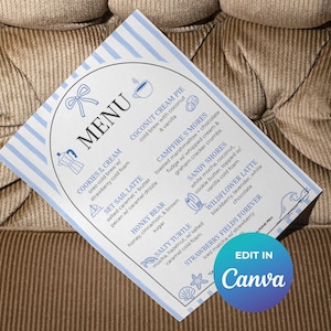Pode incluir: Um cartão de menu retangular com uma borda de listras azuis e brancas e uma moldura oval central. O menu lista bebidas de café como Cookies & Cream, Coconut Cream Pie, Campfire S'mores, Sandy Shores, Set Sail Latte, Wildflower Latte, Honey Bear, Salty Turtle e Strawberry Fields Forever, acompanhadas por pequenos ícones de linha azul de um laço, xícara de café, cafeteira, veleiro, prancha de surfe, estrela-do-mar e concha.