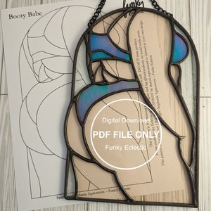 May include: A stained glass butterfly pattern with a blue, purple and teal colour scheme. The pattern is framed in black metal and is hanging from a black chain. The pattern is labelled "Booty Babe" and "Funky Eclectic".
