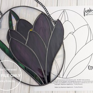 Puede incluir: Un patrón de vitral de una flor de crocus púrpura con hojas verdes. El patrón está en un círculo y está rodeado por una cadena negra. El patrón también está disponible como descarga digital.
