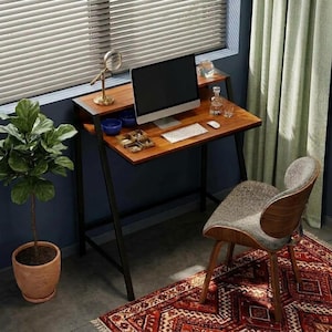 May include: A wooden desk with a black metal frame, featuring a computer monitor, keyboard, mouse, and small accessories on the surface. A curved chair with a gray fabric seat and wooden back sits in front of the desk, next to a potted plant.
