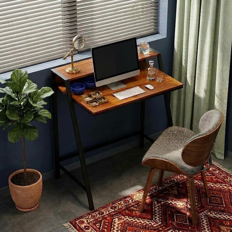 May include: A wooden desk with a black metal frame, featuring a computer monitor, keyboard, mouse, and small accessories on the surface. A curved chair with a gray fabric seat and wooden back sits in front of the desk, next to a potted plant.