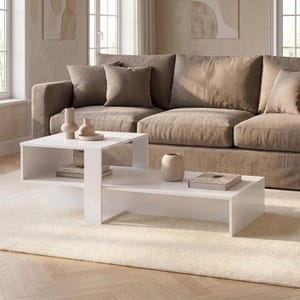 May include: A white coffee table with a multi-level design featuring an elevated square section and a lower rectangular surface. The table has open storage compartments and is constructed with clean, straight lines and a smooth, solid white finish.