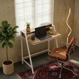 May include: A wooden desk with a white metal frame, featuring a laptop, a small potted plant, and a pen holder on the surface. A brown leather office chair with a chrome base sits in front of the desk, placed on a patterned rug near a floor lamp.