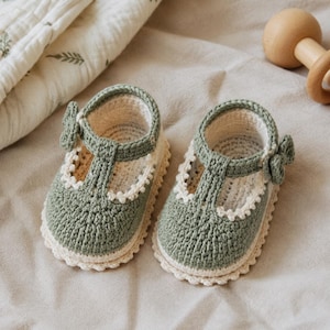 Peut inclure: Une paire de chaussures pour bébé en crochet vert sauge et crème, dotées d'une bride en T avec un petit nœud latéral, d'un bord festonné le long de l'ouverture et d'une construction en maille texturée. Elles ont un bout arrondi et une semelle épaisse.