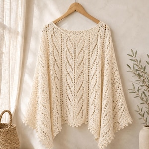 May include: A cream-colored, loose-fitting poncho featuring an intricate openwork knit pattern with vertical leaf-like motifs. The garment has a wide neckline, a draped silhouette, and a scalloped hemline with small decorative loops along the edges.