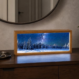May include: A rectangular light box with a wooden frame displaying a winter landscape scene. The image features snow-covered pine trees under a dark blue night sky filled with a bright, glowing Milky Way galaxy and numerous visible stars.