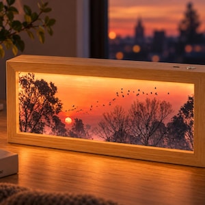 May include: A rectangular light box with a light wood frame displaying an illuminated sunset scene. The image features a bright orange sun, silhouettes of trees, and a flock of birds flying across the sky. A small power button is visible on top.