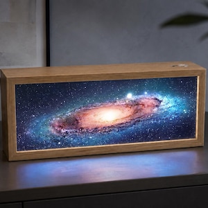 May include: A rectangular light box with a light-coloured wooden frame featuring a vibrant, illuminated image of a spiral galaxy. The galaxy display shows a bright central core with swirling blue and pink nebulae against a dark starry space background.