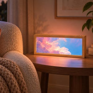 May include: A rectangular light box with a light-coloured wooden frame displaying a vibrant, illuminated image of a sunset sky filled with pink, orange, and blue clouds. The device emits a soft, warm glow, casting light onto the surrounding area.