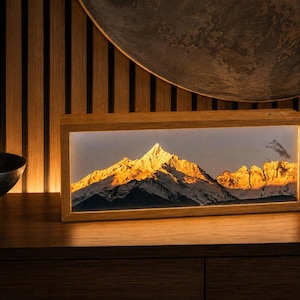 May include: A rectangular light box with a wooden frame displaying a backlit photographic print of a snow-capped mountain peak illuminated by golden sunlight. The scene features a sharp, triangular summit with rocky slopes and deep shadows.