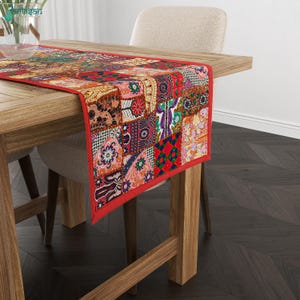 May include: A rectangular table runner featuring a vibrant patchwork design of various colorful fabric squares with intricate floral and geometric patterns, finished with a solid red fabric border along the edges of the textile piece.