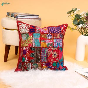 May include: A square throw cushion featuring a vibrant patchwork design of red, pink, blue, and gold fabric squares. The surface is decorated with intricate embroidery, floral patterns, and small reflective mirror accents stitched into the fabric.