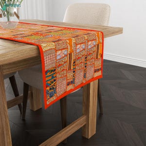 May include: A rectangular table runner featuring a vibrant patchwork design with various orange, red, and gold fabric panels. The runner displays intricate floral and geometric patterns, finished with a solid orange border along the edges.