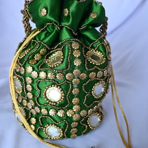 May include: A green satin drawstring pouch bag featuring gold-toned beadwork, small metallic flower-shaped embellishments, and circular mirror accents arranged in a geometric pattern. The bag includes a gold cord closure with metallic bell-shaped ends.