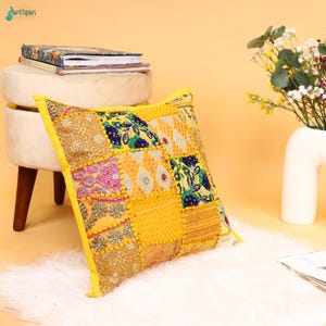 Puede incluir: Un cojín decorativo cuadrado con un diseño de patchwork de varios tejidos en amarillo, dorado, rosa y azul. El cojín está adornado con intrincados bordados, lentejuelas y detalles de hilo metálico, rematado con un borde amarillo sólido.