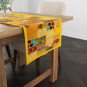 May include: A yellow table runner featuring a patchwork design of embroidered fabric squares in various colors including red, blue, green, and gold. The runner displays intricate floral and geometric patterns across its rectangular surface.