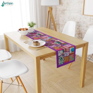 May include: A rectangular table runner featuring a vibrant patchwork design with various floral and geometric patterns in shades of purple, pink, orange, and green, centered on a light wood dining table with a white plate and a bowl of fruit.