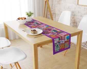 May include: A rectangular table runner featuring a vibrant patchwork design with various floral and geometric patterns in shades of purple, pink, orange, and green, centered on a light wood dining table with a white plate and a bowl of fruit.