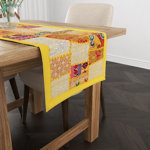 May include: A rectangular table runner featuring a patchwork design of various embroidered fabric squares in yellow, red, orange, and white. The runner has a solid yellow border and displays intricate floral and geometric patterns throughout.