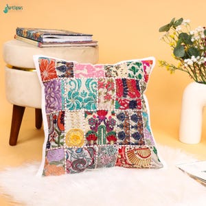 Könnte beinhalten: Ein quadratisches Zierkissen mit einem Patchwork-Design aus verschiedenen bestickten Stoffquadraten in leuchtenden Farben wie Rot, Blau, Grün, Gelb und Violett. Das Kissen hat einen weißen Rand und zeigt komplexe florale und geometrische Muster.