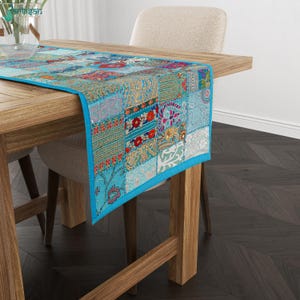 May include: A rectangular table runner featuring a vibrant patchwork design with various blue, red, and gold floral and geometric patterns. The fabric is finished with a solid bright blue border and is draped over a wooden dining table.
